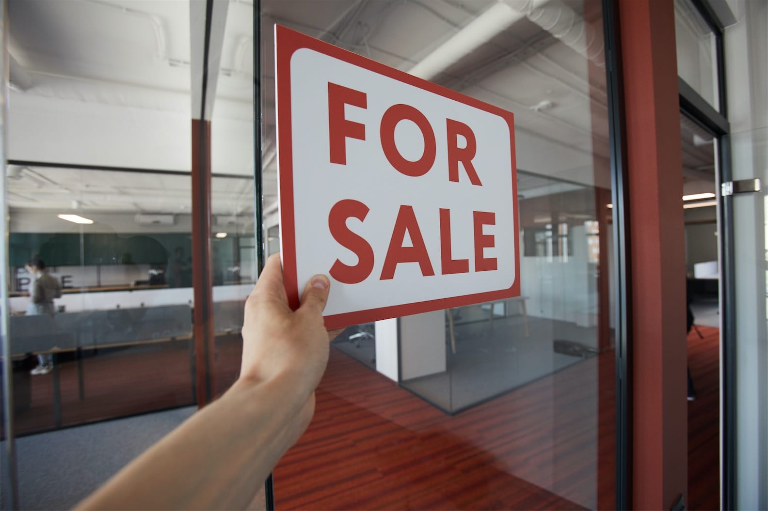 A hand holds a "For Sale" sign near a glass door inside a modern office space, hinting at possible business transformation services. The office, featuring wooden flooring and glass walls, has visible workstations in the background, perfectly suited for part-time CFO services in Southeast Queensland.