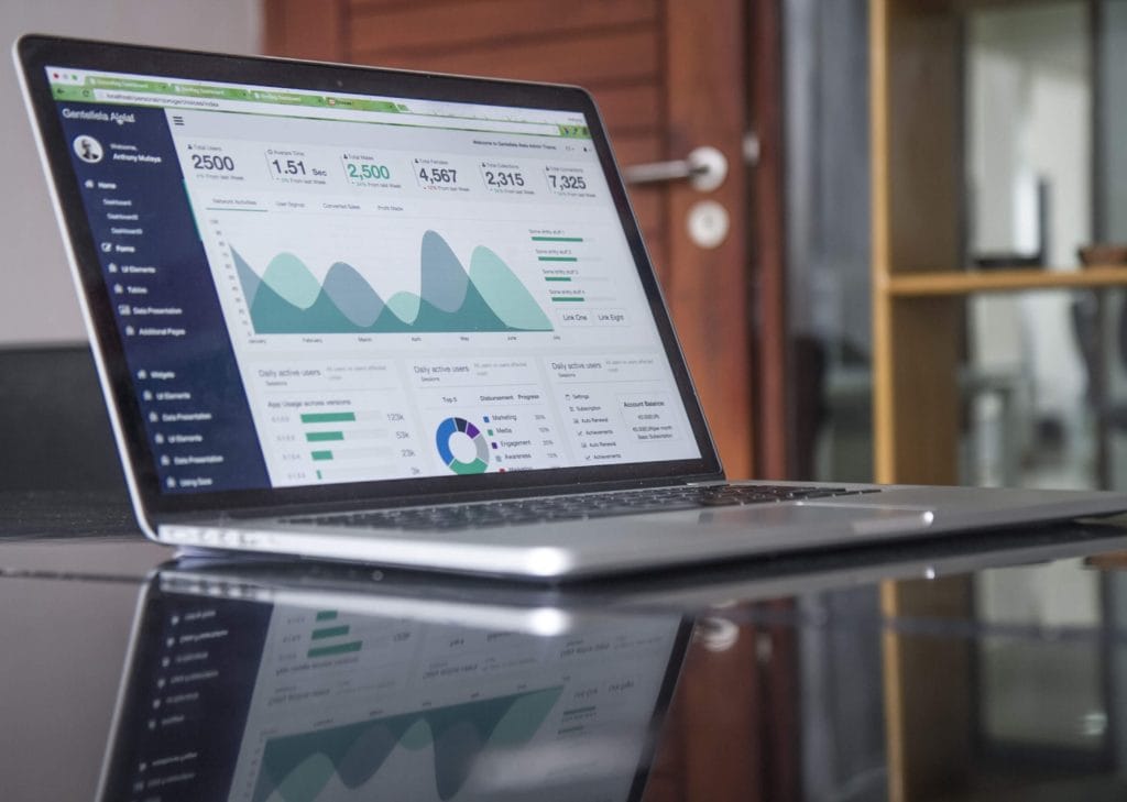 A laptop on a glass table displays a dashboard with various graphs and charts, illustrating data analytics for virtual CFO services Australia. The background features a wooden cabinet and an open door.